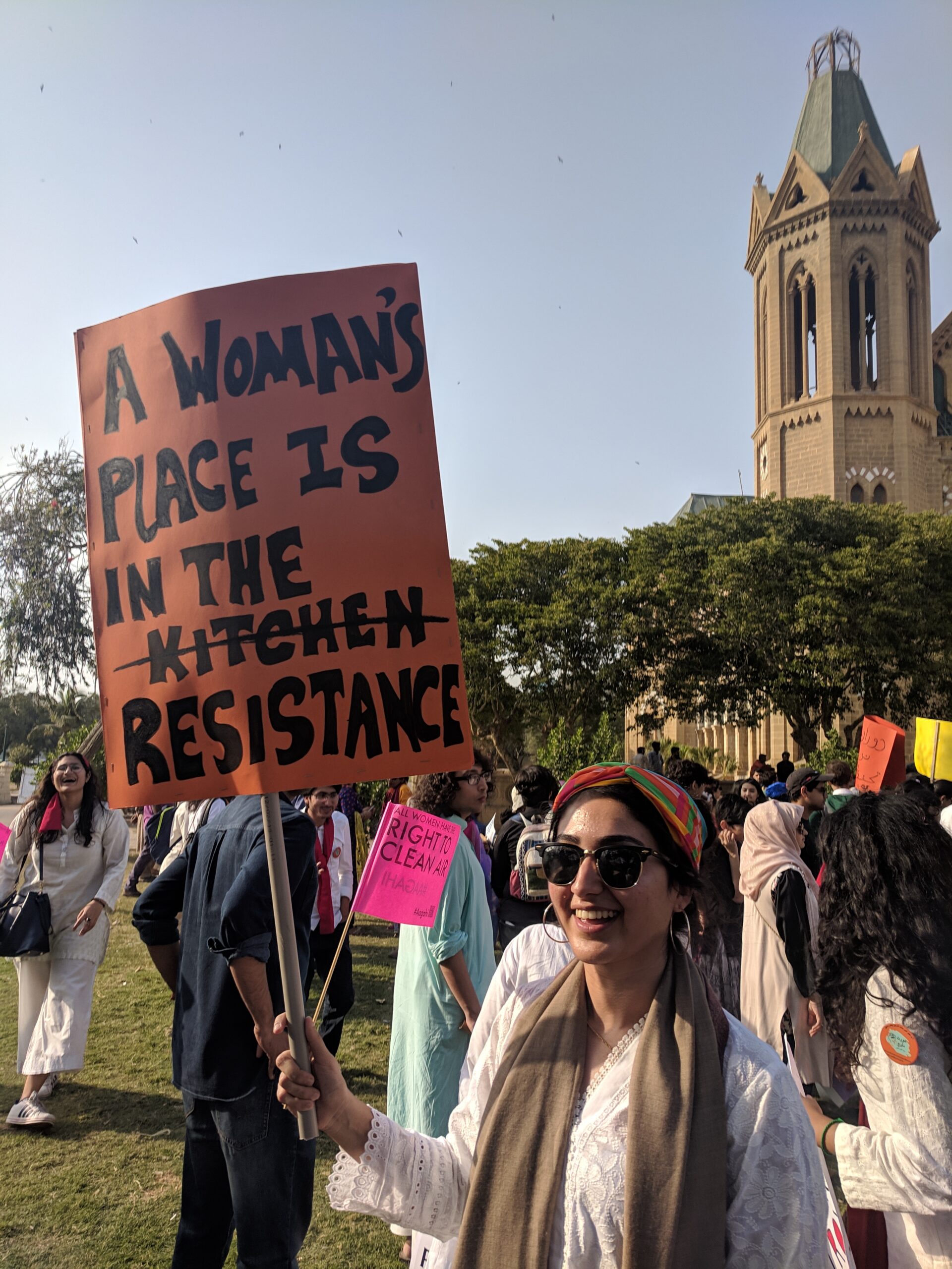 A smiling woman holding an orange placard that reads "A woman's place is in the k̶i̶t̶c̶h̶e̶n̶  resistance"