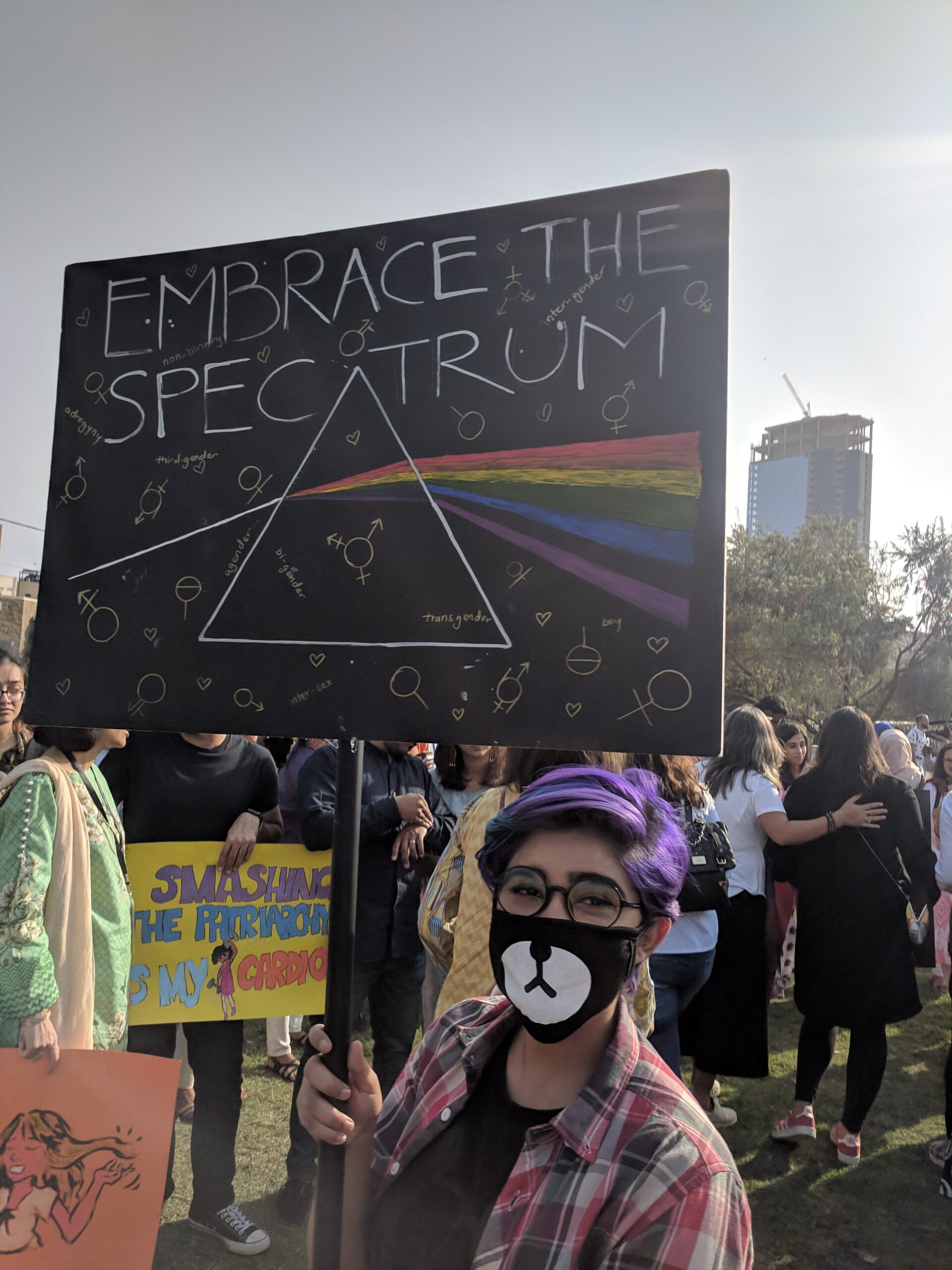 A person with purple hair, wearing a mask, holds a sign that reads "Embrace the spectrum". It has an illustration of light passing through a triangular prism to form a rainbow