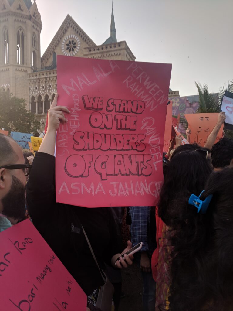 A person holding a pink sign that reads "We stand on the shouldrs of giants", with the words surrounded names of promiment feminist such as Malala, Perween Rehman, Asma Jahangir, Sabeen Mehmud, and Qandeel Baloch