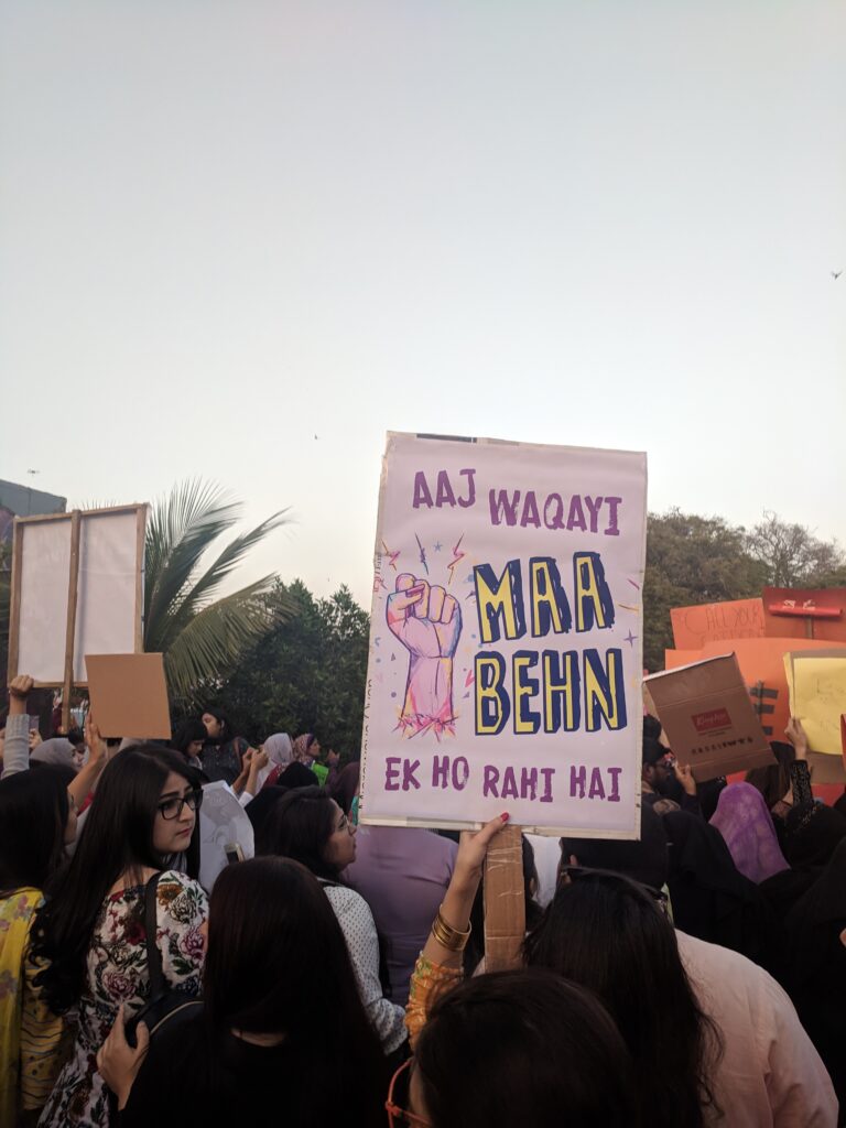 A sign held up in the midst of a crowd reads, "Aaj waqayi Maa Behn ek ho rahi hai", with an illustrated fist adjacent to the words